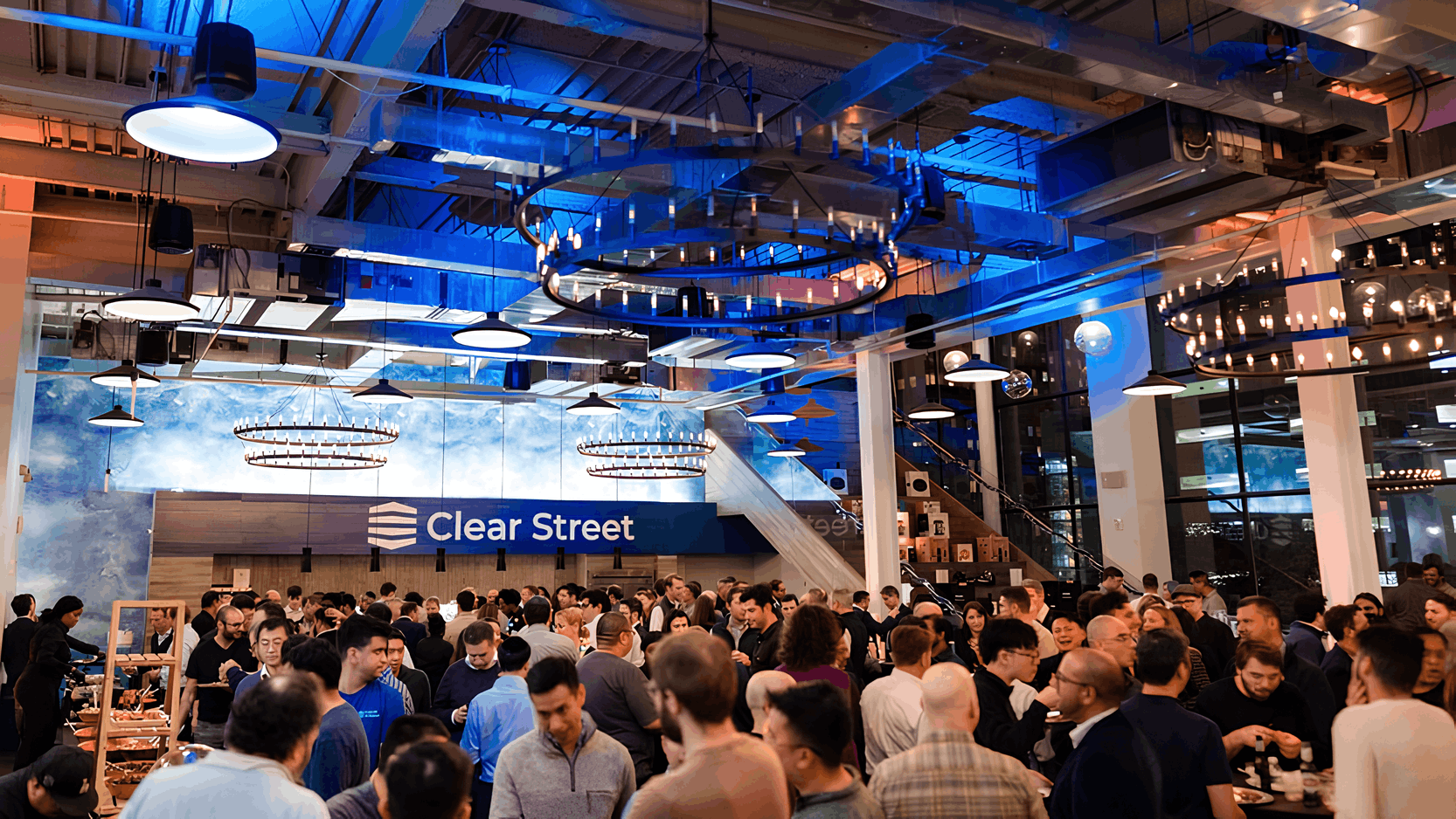 Crowded networking event at Clear Street office with blue and orange lighting, modern decor, and large chandeliers.