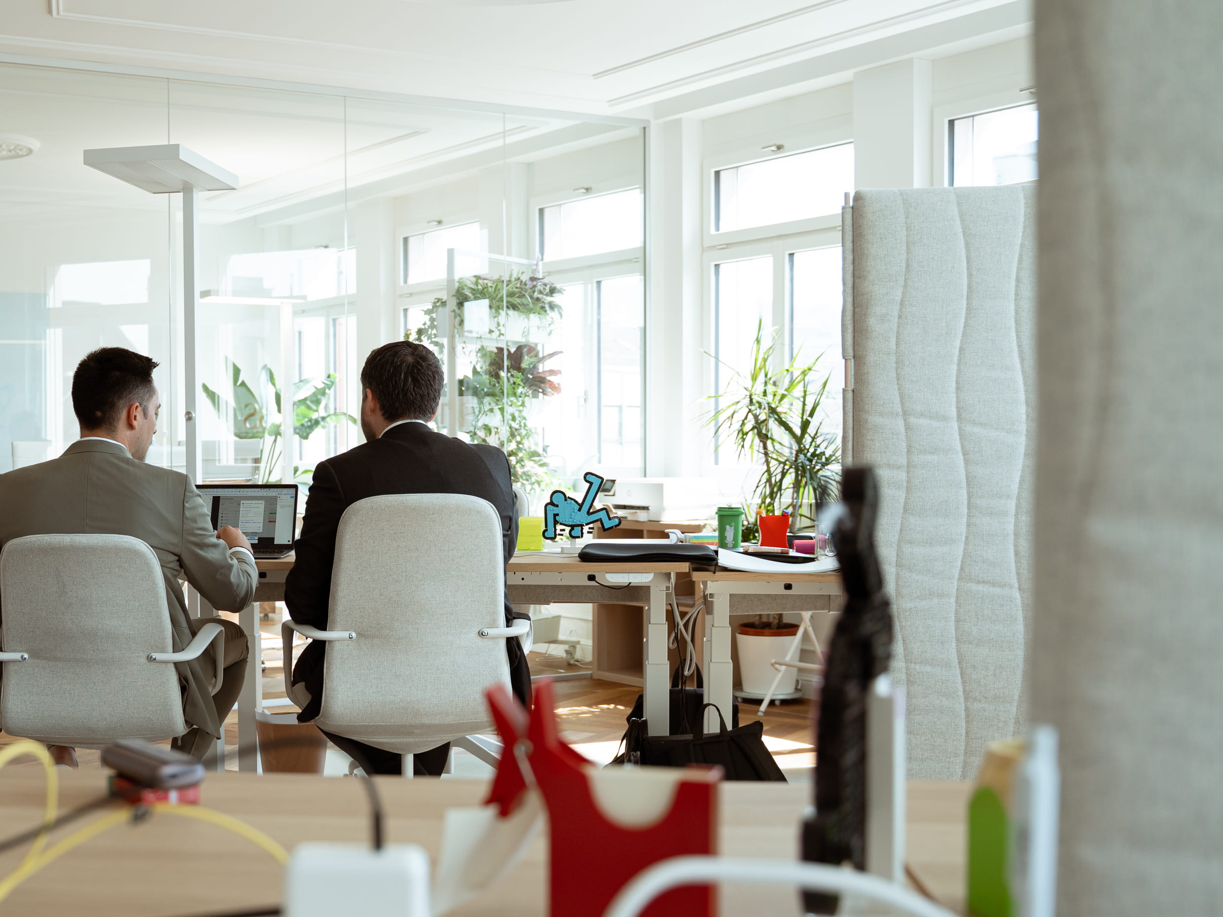 Two men collaborating in a modern, bright office with plants and computers.