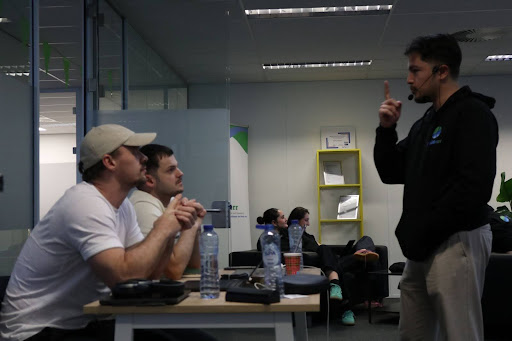 Group discussion in an office, with three people seated at a table and one standing, using a microphone headset.