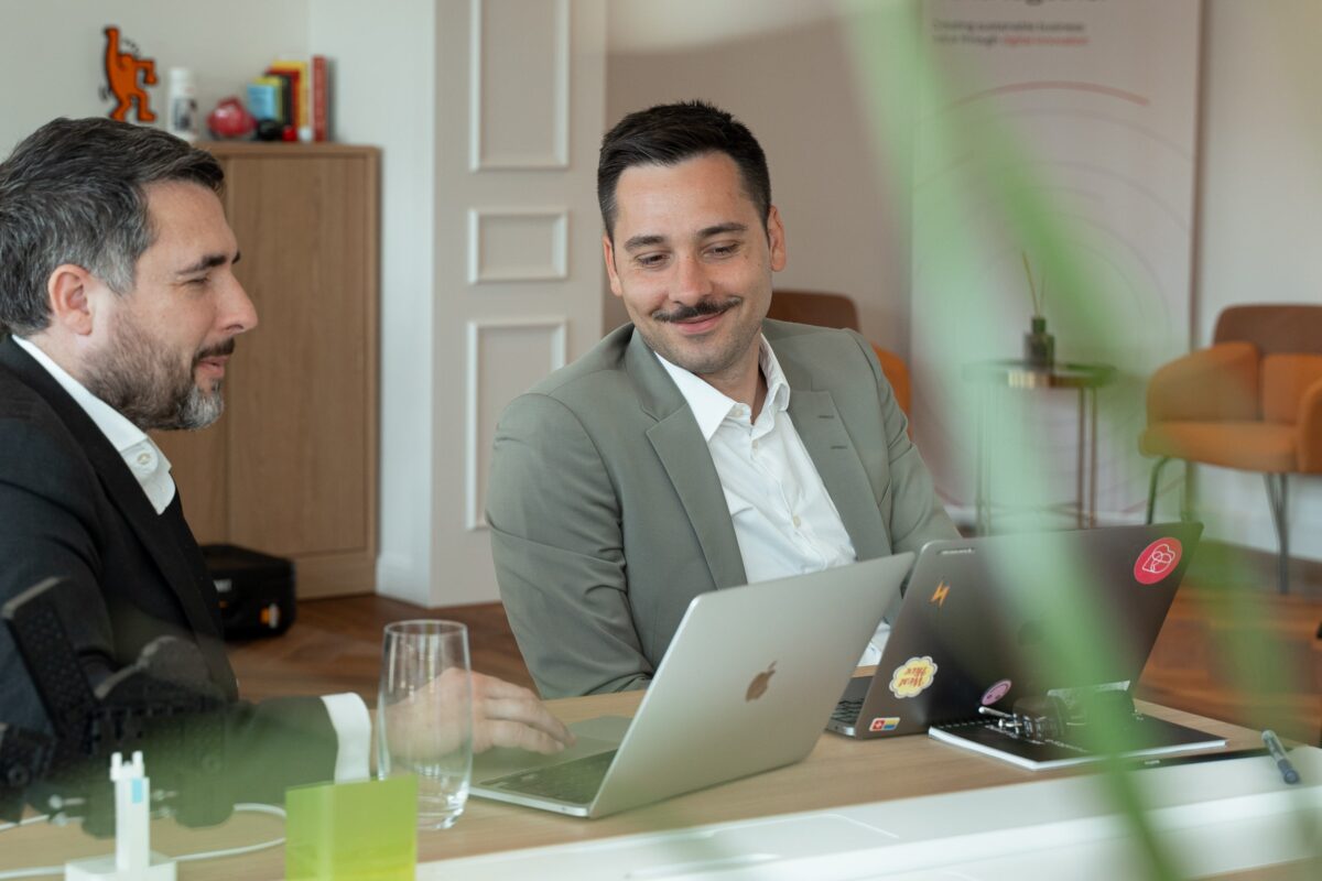 Two businessmen collaborating on laptops in a modern office setting.