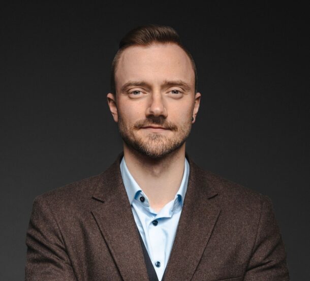 Confident man in brown blazer and blue shirt against a dark background, looking directly at the camera.