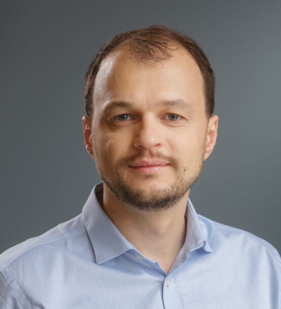 Portrait of a man in a light blue shirt against a gray background, looking at the camera with a neutral expression.