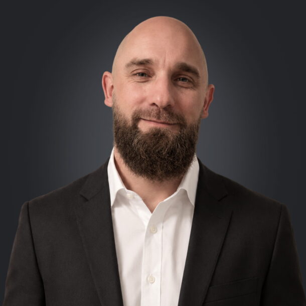 Bald man with a beard in a suit smiling against a dark background.