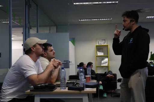 Group discussion in an office, with three people seated at a table and one standing, using a microphone headset.