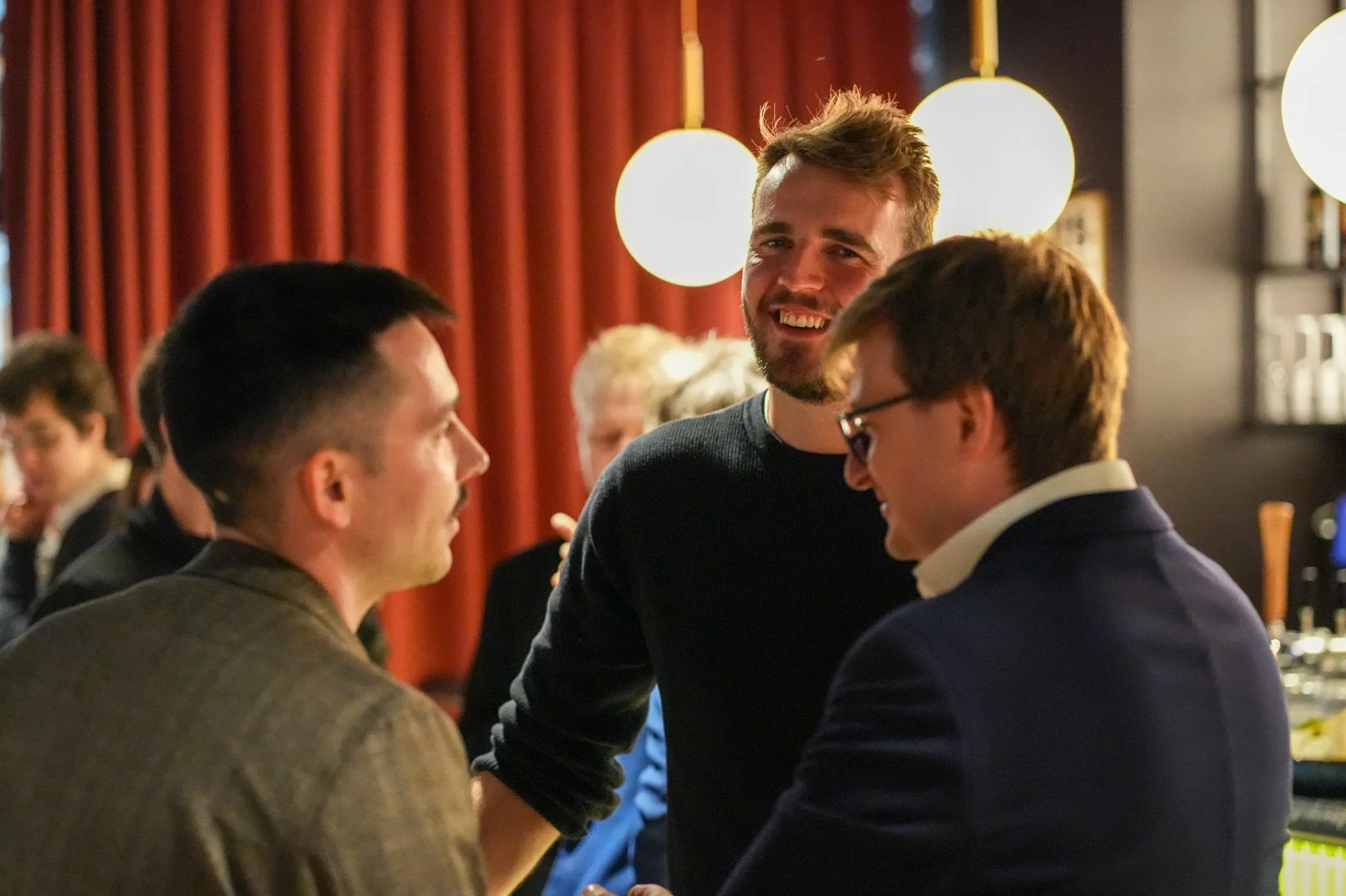 Group of people enjoying conversation at a social event with warm lighting and red curtains in the background.