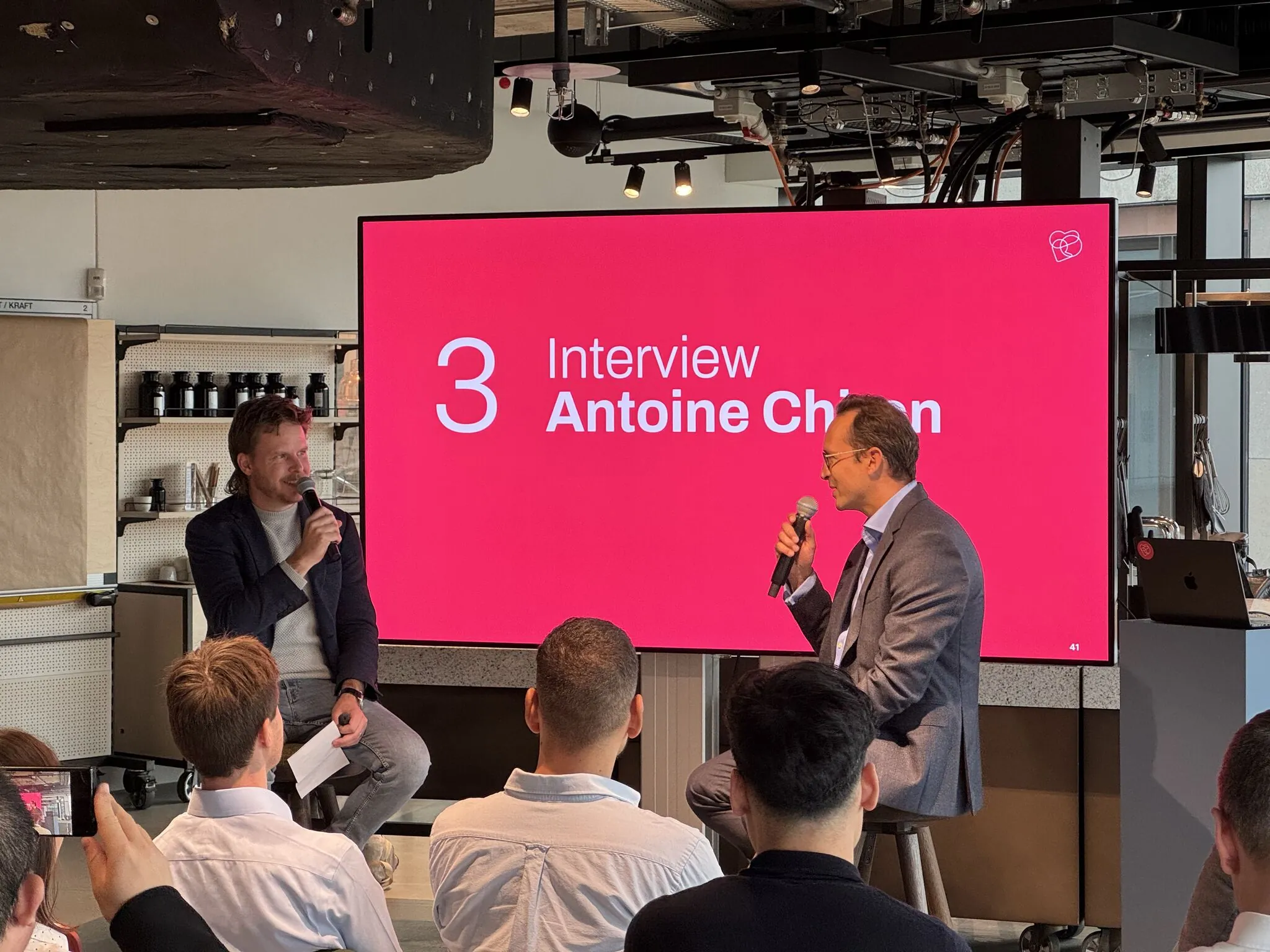 Two people conducting an interview on stage with an audience, large screen displays text in the background.