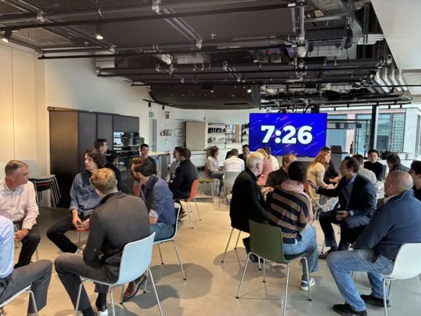 People in a modern office setting engaged in group discussions, large digital clock displaying 7:26 in the background.