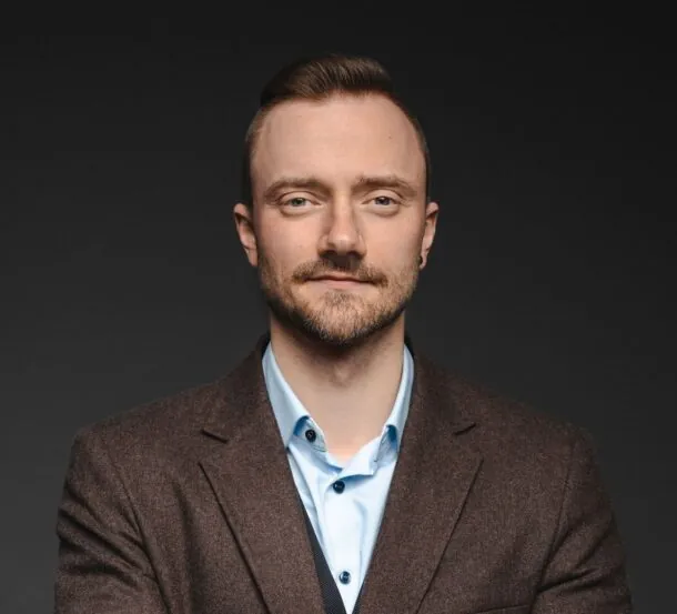 Confident man in brown blazer and blue shirt against a dark background, looking directly at the camera.