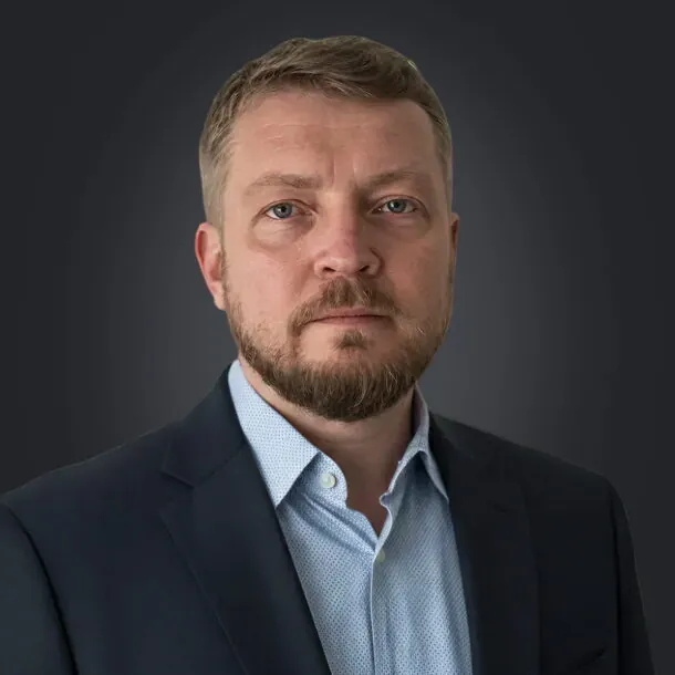 A bearded man in a suit and light blue shirt against a dark background.