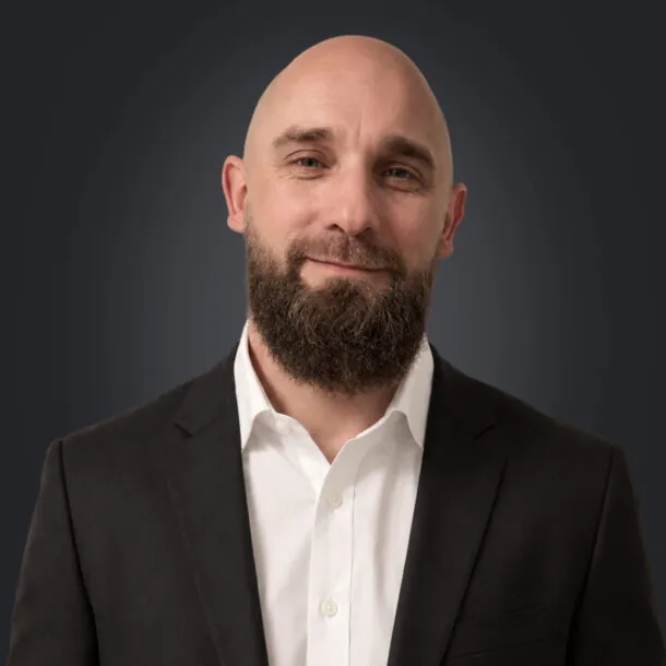 Bald man with a beard in a suit smiling against a dark background.